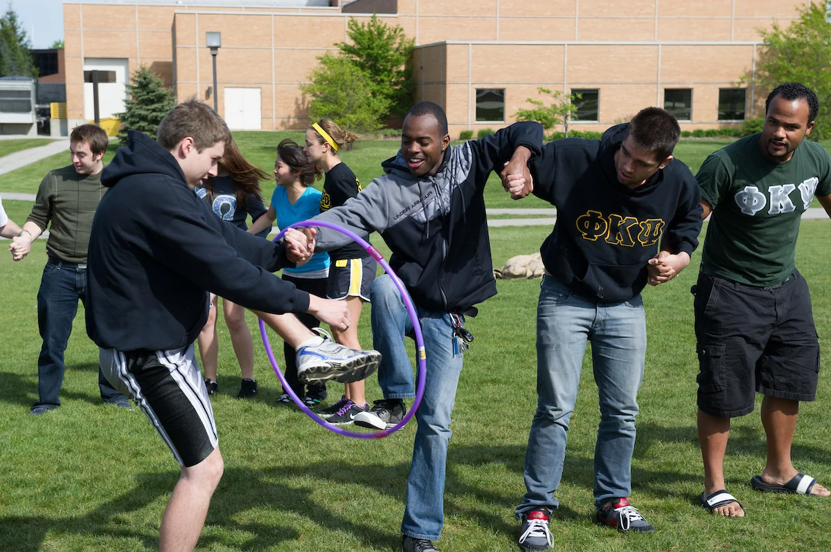 Students, including International Academics, pass a hula hoop along a line while holding hands on a grassy campus lawn.