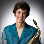 Stacy Maugans, with short brown hair and glasses, smiles while holding a saxophone and wearing a teal sheer top.