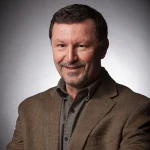 A man in a brown blazer smiling, with short dark hair and a goatee, posing against a gray background.