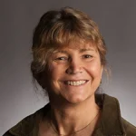Smiling woman with light brown hair, wearing a green jacket, in front of a neutral gray background—Elizabeth Gingerich, JD.