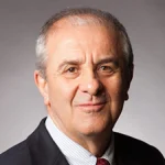 Musa Pinar, Ph.D., an older man in a suit and tie, smiles warmly at the camera against a plain background.