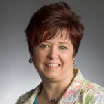 Smiling woman with short reddish-brown hair, identified as Kelly Migler, wearing a patterned jacket and blue top against a gray background.
