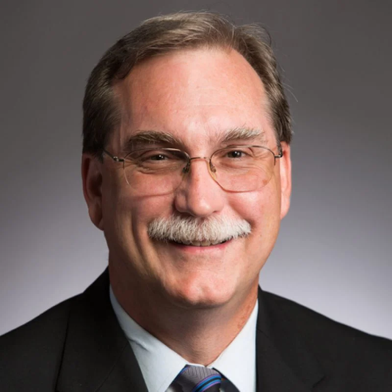 Smiling middle-aged man, Rick Gillman, with glasses, mustache, and a suit against a plain gray background.