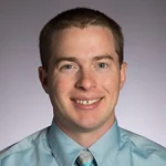 A man with short brown hair smiles, wearing a light blue shirt and striped tie against a plain background—Luke Venstrom, Ph.D.