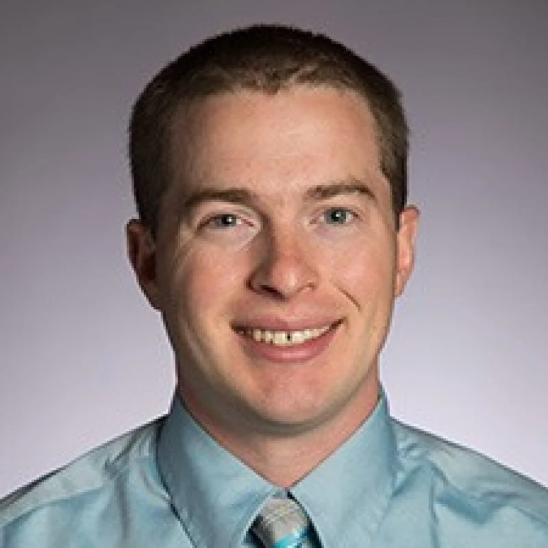 A man with short brown hair smiles, wearing a light blue shirt and striped tie against a plain background—Luke Venstrom, Ph.D.