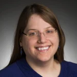 Smiling Lara Pudwell with glasses, long brown hair, and a blue sweater, against a dark gradient background.
