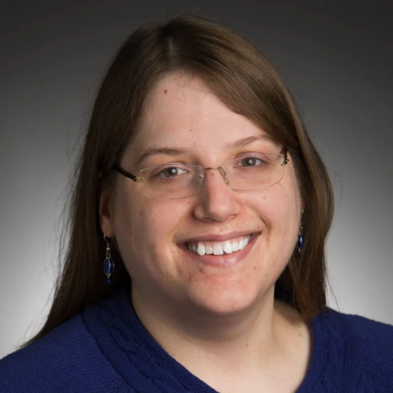 Smiling Lara Pudwell with glasses, long brown hair, and a blue sweater, against a dark gradient background.