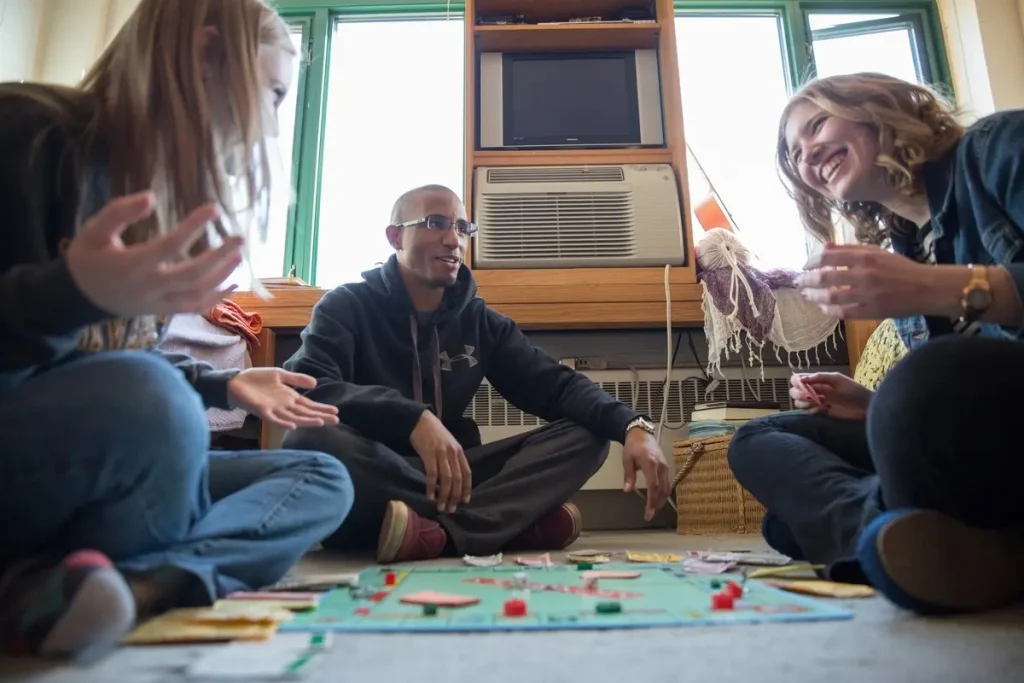 Three friends sit on the floor of their off-campus housing, smiling and playing a board game together near a window.