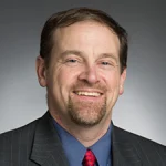 Smiling man in a suit, blue shirt, and red tie against a gray background—Jeffrey Will, Ph.D.