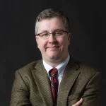 Edward Upton, a man in glasses wearing a brown plaid blazer, white shirt, and striped tie, smiles confidently with his arms crossed.
