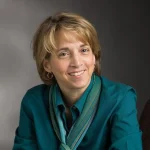 Gretchen Buggeln, a woman with short blonde hair in a teal shirt and scarf, smiles at the camera against a gray background.