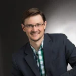 Samuel Graber, a man with glasses and short brown hair, wears a blazer and plaid shirt, smiling in front of a gray background.