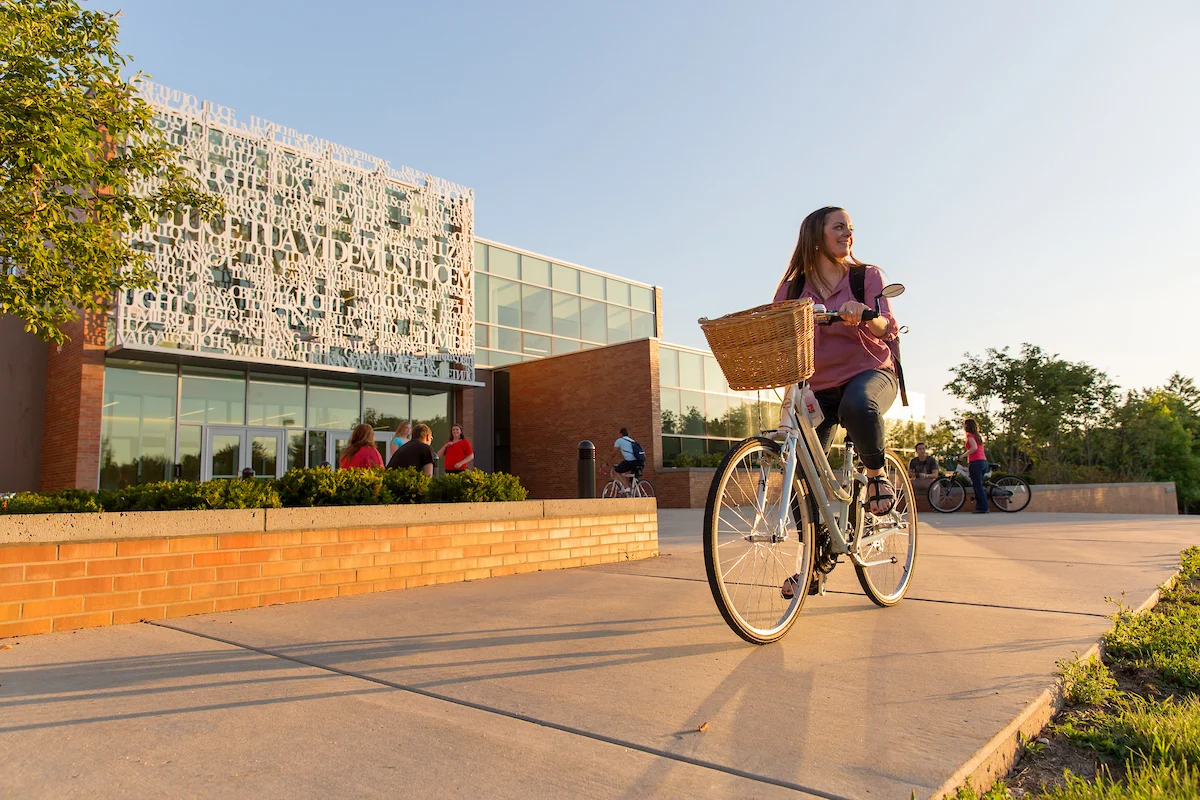 A woman rides a bicycle with a basket past parking spaces on a sunny sidewalk outside a modern campus building.