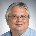 Middle-aged man, Michael K. Watters, with gray hair, glasses, and a blue shirt, smiling at the camera against a gray background.