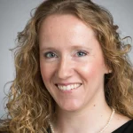Anne Marie Bice, a woman with curly blonde hair, smiles warmly while wearing a black top and pearl necklace against a neutral background.