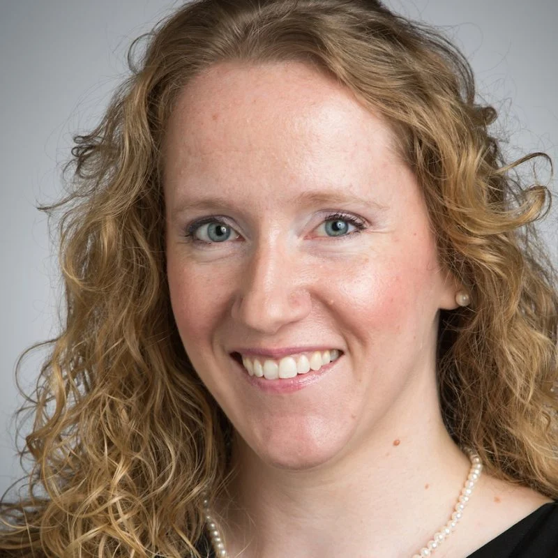 Anne Marie Bice, a woman with curly blonde hair, smiles warmly while wearing a black top and pearl necklace against a neutral background.