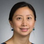 Jiangxia Liu, Ph.D., with long dark hair pulled back and blue earrings, smiles warmly at the camera against a gray background.