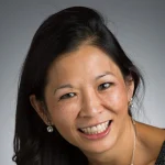 Smiling woman with long dark hair, wearing earrings and a necklace, against a gray background.