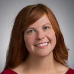 Woman with long red hair smiling, wearing a red top, photographed against a plain grey background. Mandy Morrill-Richards, LMHC, NCC, exudes warmth and professionalism in her approachable portrait.
