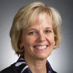 Smiling woman with short blonde hair, wearing a dark jacket and patterned blouse, on a gray background. Julie Peller, PhD.