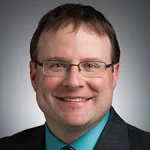 Smiling man, identified as Robert W. Clark, with glasses in a suit and teal shirt, posing in front of a gray background.