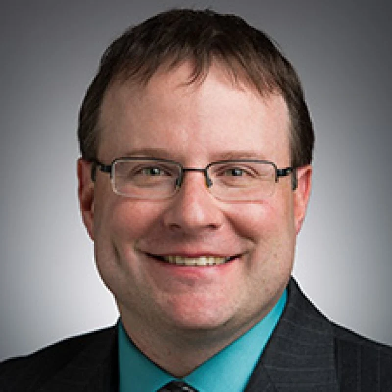 Smiling man, identified as Robert W. Clark, with glasses in a suit and teal shirt, posing in front of a gray background.