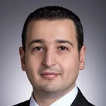 Georges El-Howayek, Ph.D., in a suit and tie, poses for a professional headshot against a gray background.