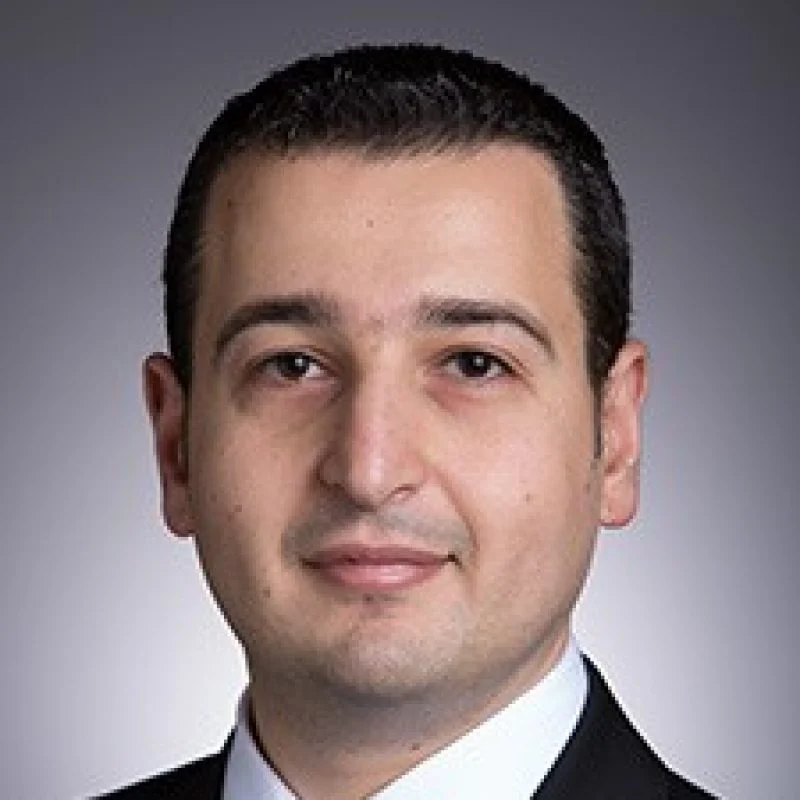 Georges El-Howayek, Ph.D., in a suit and tie, poses for a professional headshot against a gray background.