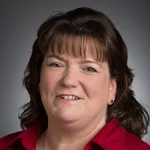 Laura Sanders, a woman with brown hair and bangs, smiles warmly in a red collared shirt against a gray background.