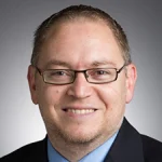 Smiling man with glasses, short hair, and a suit jacket, Sami Khorbotly, Ph.D., stands in front of a neutral gray background.