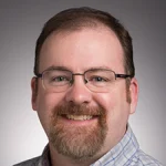 Smiling man with glasses, short brown hair, and beard, wearing a checked shirt in front of a gray background—Michael Longan, PhD.