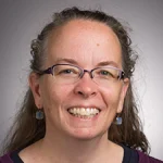 Smiling woman with glasses and long wavy hair, wearing earrings and a dark top, against a gray background—Dr. Teresa Bals-Elsholz, Ph.D.