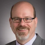 Smiling man with glasses, goatee, and suit against a neutral gray background—Andrew Richter exudes confidence and professionalism.