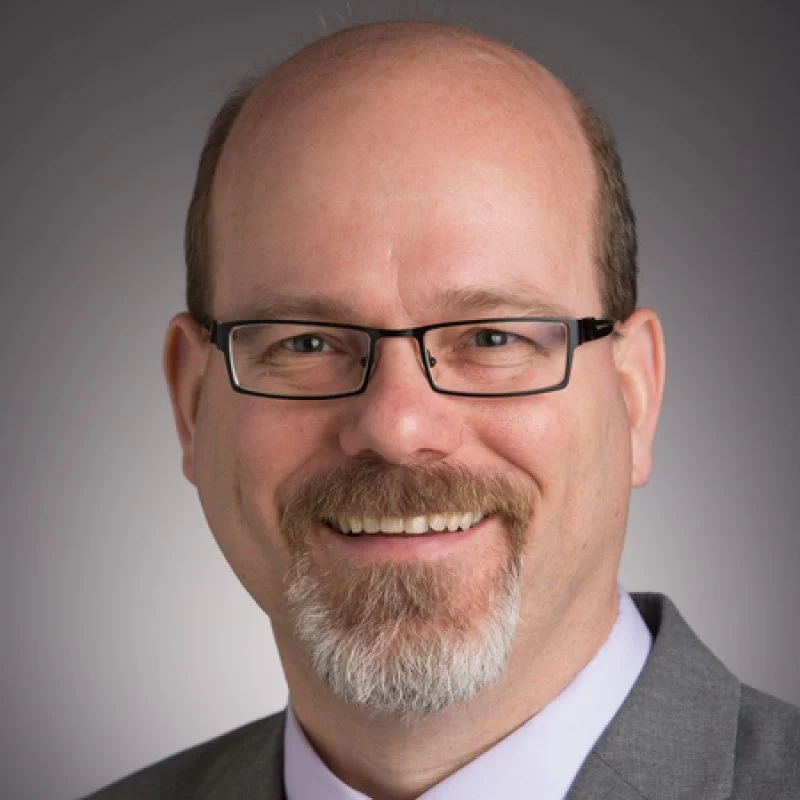 Smiling man with glasses, goatee, and suit against a neutral gray background—Andrew Richter exudes confidence and professionalism.