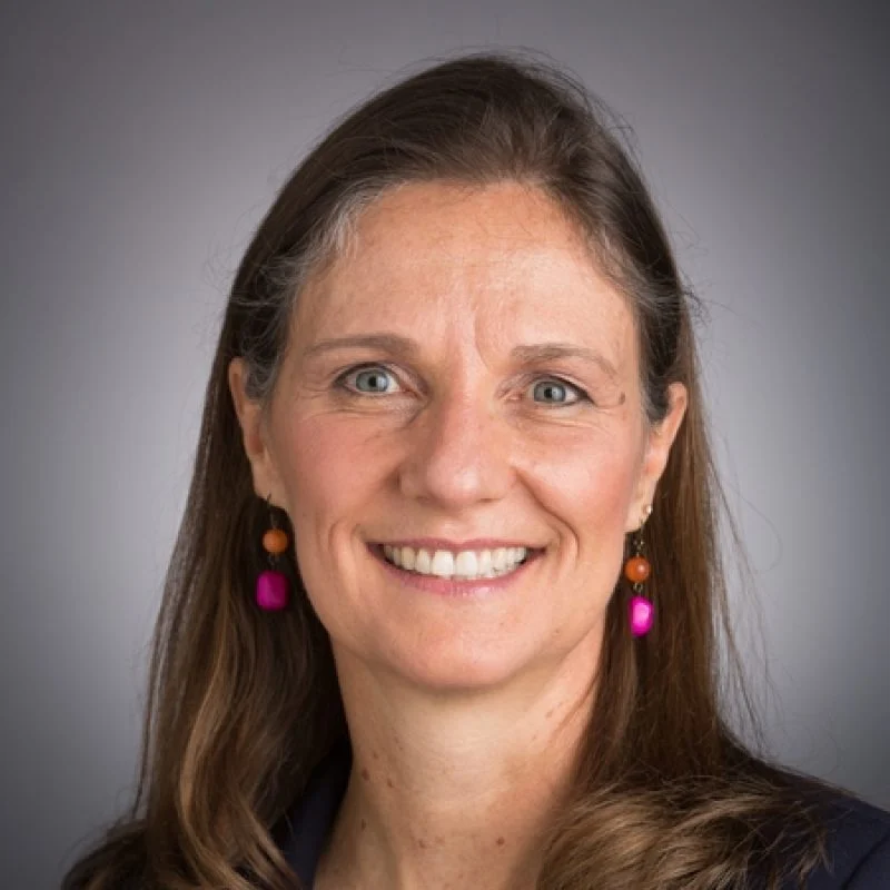 Smiling woman with long brown hair, wearing pink earrings, against a plain gray background—Barbara Crumpacker Niedner.