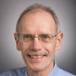 Smiling older man with glasses, short light hair, and a mustache, wearing a light blue collared shirt—Bruce Hrivnak.