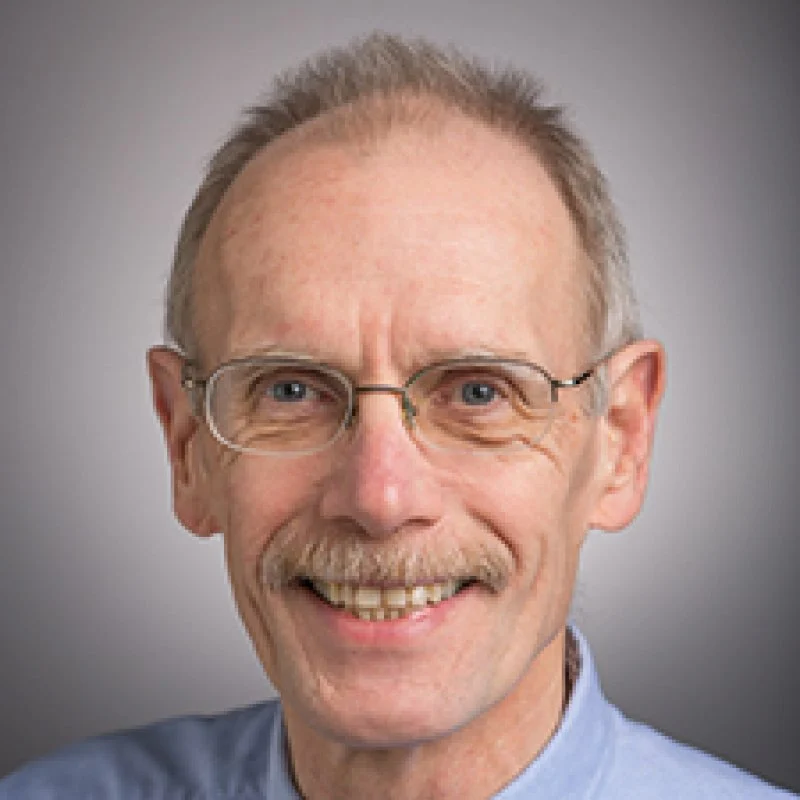 Smiling older man with glasses, short light hair, and a mustache, wearing a light blue collared shirt—Bruce Hrivnak.