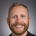 Smiling man with short blond hair and beard, wearing a dark shirt, in front of a gray background—Peter Johnson, Ph.D.