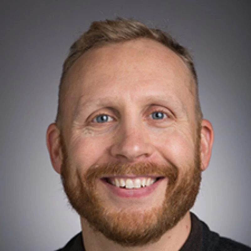 Smiling man with short blond hair and beard, wearing a dark shirt, in front of a gray background—Peter Johnson, Ph.D.