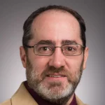 Todd Hillwig, a man with glasses, short brown hair, and a beard, wears a tan jacket and maroon shirt while smiling slightly.