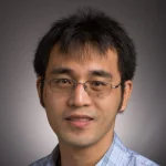A man with short black hair, glasses, and a plaid shirt, smiling at the camera against a gray background.