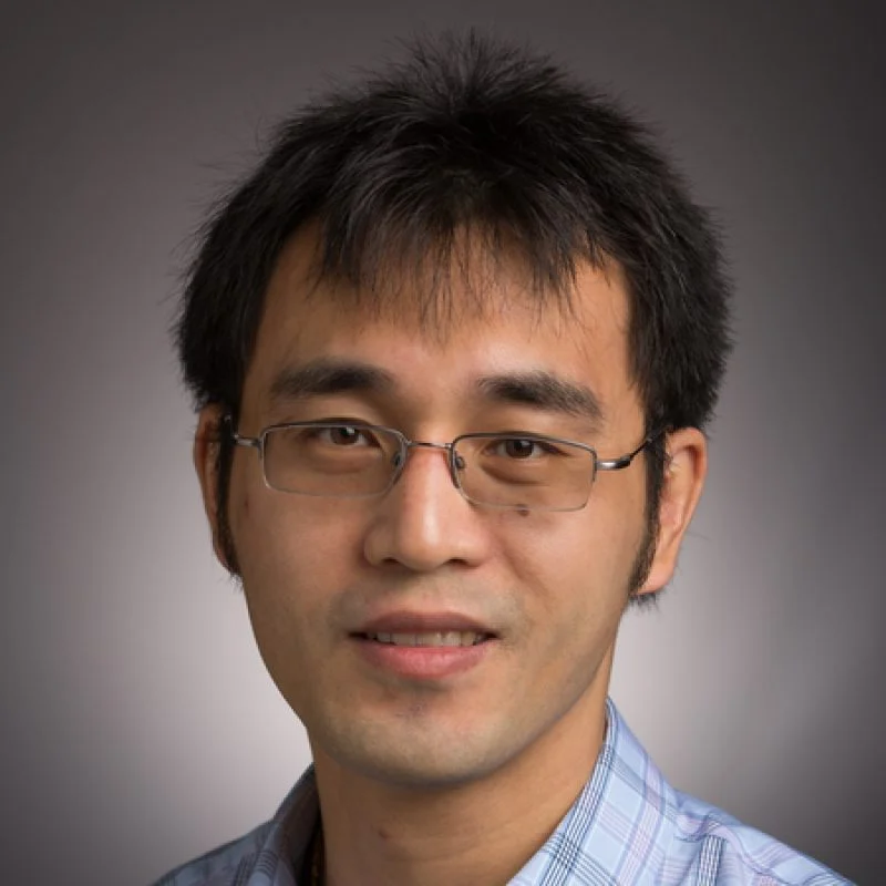 A man with short black hair, glasses, and a plaid shirt, smiling at the camera against a gray background.