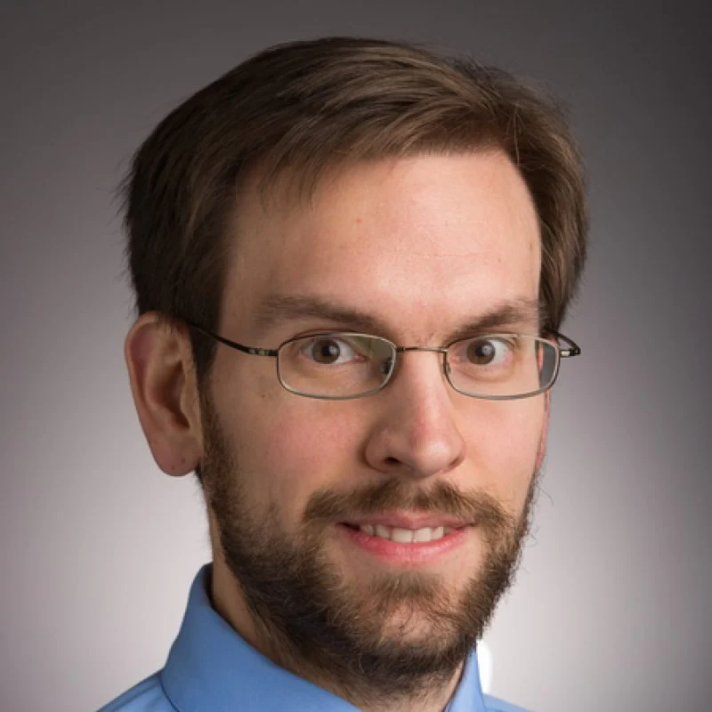 A man with glasses, short brown hair, and a beard, wearing a blue collared shirt, smiling slightly.