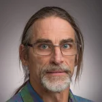 Middle-aged man with glasses, long hair, a mustache, and a beard, wearing a plaid shirt, stands against a gray background—Patrick Sullivan.