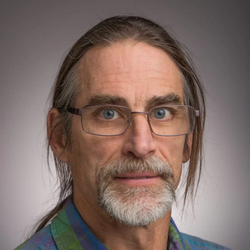 Middle-aged man with glasses, long hair, a mustache, and a beard, wearing a plaid shirt, stands against a gray background—Patrick Sullivan.