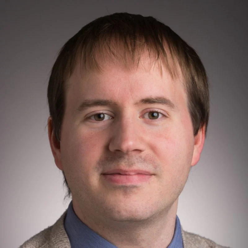 A man with straight brown hair, light skin, and a slight smile wearing a blue shirt and beige sweater.