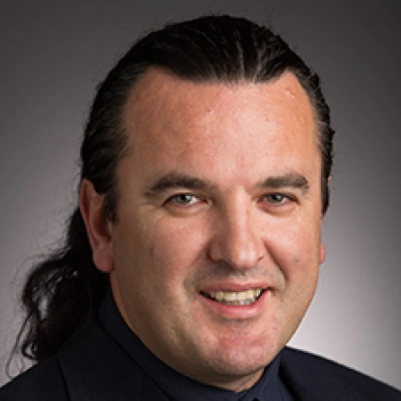 Smiling man with dark hair pulled back, wearing a dark suit, stands against a dark background—Saso Poposki exudes confidence and style.