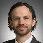 Adam Gibson-Even, a man with short curly hair, beard, and mustache, is wearing a suit and smiling at the camera.
