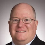 Smiling man with glasses, bald head, and suit jacket stands before a neutral background—Carmine Polito, Ph.D., exudes professionalism and warmth.