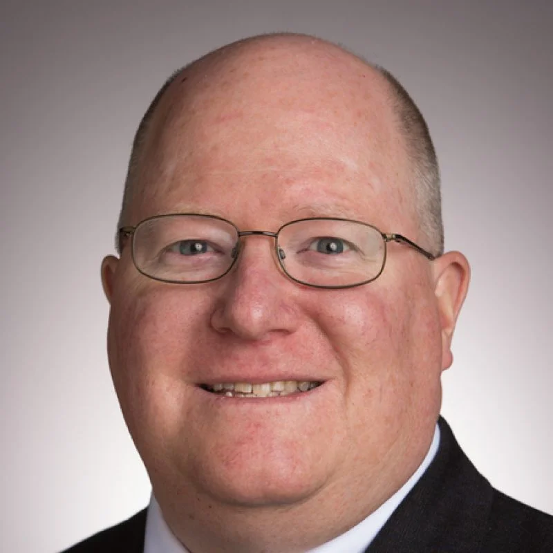 Smiling man with glasses, bald head, and suit jacket stands before a neutral background—Carmine Polito, Ph.D., exudes professionalism and warmth.